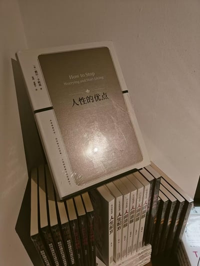 这是一张竖版实拍照片，画面呈现了一摞堆叠放置的励志、心理类书籍。最上方是戴尔·卡耐基的经典著作《人性的优点》（英文原名*How to Stop Worrying and Start Living*），书籍带有塑封，为全新状态。下方整齐排列着多本重复的同类型书籍，包括《如何在博弈和沟通中获得更多》《5分钟漫画人生配方》《人间清醒》等，同样带有塑封。场景位于室内浅色墙面的角落，整体氛围偏向沉静的阅读相关场景。