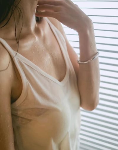 图片展示了一位女性的上半身特写，她穿着一件浅色V领背心，皮肤上有明显的水珠，暗示刚沐浴或接触过水。她的右手轻触颈部，左臂佩戴着一个银色手镯。背景是带有水平条纹的百叶窗，光线透过百叶窗在皮肤上形成了柔和的光影效果。整体画面呈现出一种清新、略带湿润的氛围，突出了肌肤质感和衣物的轻薄特性。