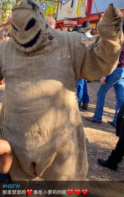 图片中展示了一位身穿粗麻布稻草人服装的人，服装面部有缝线制成的诡异微笑和空洞的黑色眼洞。人物站在户外场地中，背景有其他游客和彩色摊位，推测是在万圣节活动或嘉年华现场。人物右手举起，做出类似打招呼的姿势。图片底部有明显的中文文字覆盖：“#NSFW 那里瑟瑟的❤️像是小萝莉的呢❤️❤️❤️”，内容涉及不当联想，将成人服装与儿童形象进行不适当关联。