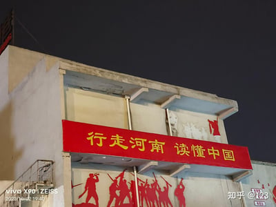 夜晚拍摄的建筑外景，画面中一栋浅色建筑的上部悬挂着醒目的红色横幅，横幅上用黄色字体写着“行走河南 读懂中国”。横幅下方的墙面上有红色的人物剪影图案，似表现历史或革命场景。左下角有水印“VIVO X9 | ZEISS 2023/10/02 19:40”，右下角有“知乎 @123”水印，显示拍摄设备、时间及发布平台信息。