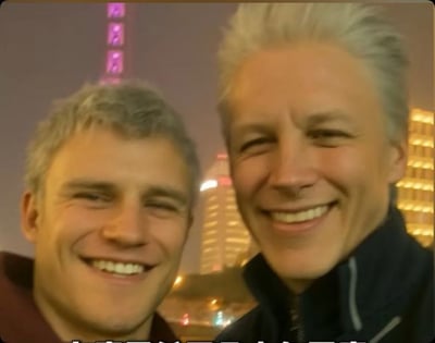 两位头发花白的男子在夜晚的上海背景下自拍，身后可见标志性的紫色灯光塔楼。两人都面带微笑，看起来心情愉悦。