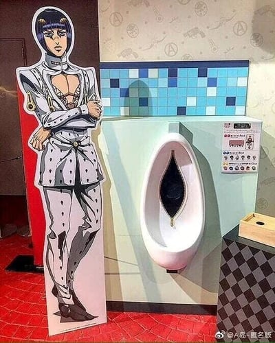 这是一张在男卫生间内拍摄的实拍图，画面右侧是一个造型独特的男用小便池，其开口的狭长形状与《JOJO的奇妙冒险：黄金之风》中角色布鲁诺·布加拉提的标志性领口拉链设计高度相似；画面左侧放置了布鲁诺·布加拉提的等身卡通立牌，他身着标志性的白色带点纹制服，双臂交叉抱于胸前，表情严肃，立牌的位置与小便池形成了巧妙的趣味呼应，利用角色的经典造型和小便池的外形制造了玩梗的效果，能让熟悉该ACG作品的观众立刻领会其中的趣味点，属于线下联动活动中设置的趣味场景。
