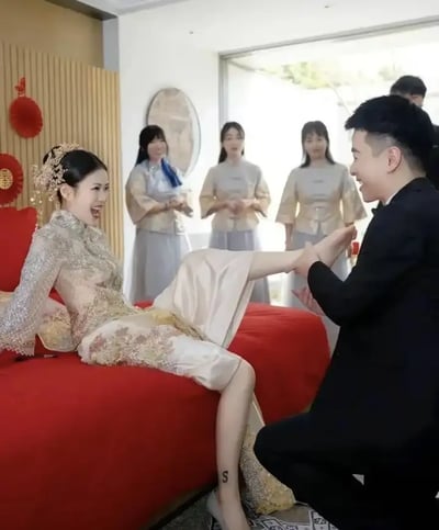 图片展示一对身着中式婚礼服饰的新人，场景明显为婚礼习俗环节。新娘坐在覆盖红色床单的大床上，腿上有一个“S”形的贴纸或纹身。新郎单膝跪地为新娘穿鞋，旁边站着几位伴娘。整体氛围温馨轻松，画面带有轻微的喜剧色彩，常见于中国婚礼表情包中。特写腿上的“S”字母有玩梗的潜力，会让网友联想到一些谐音或梗。
