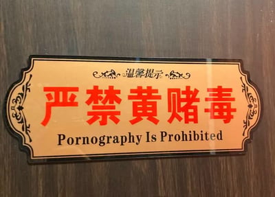 整张图是一块安装在深棕色木质墙面的公共提示标识实拍图，标识采用仿复古牌匾设计，浅橘黄色底配黑色描边与花纹装饰，顶部印有“温馨提示”字样，主体是醒目的红色大号字体“严禁黄赌毒”，但下方对应的英文翻译仅标注了“Pornography Is Prohibited”，仅翻译了“黄”对应的禁止内容，完全遗漏了赌博、毒品相关的禁止要求，属于典型的翻译疏漏错误，形成了荒诞的反差效果。