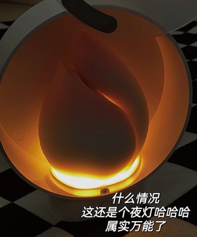 图片近距离拍摄了一个圆形设备的内部，内部呈现温暖的橙色灯光，造型类似火焰，设备放置在黑白格子图案的表面上。图片底部配有文字：“什么情况 这还是个夜灯哈哈哈 属实万能了”，表达了对该设备兼具夜灯功能的惊喜和赞叹。