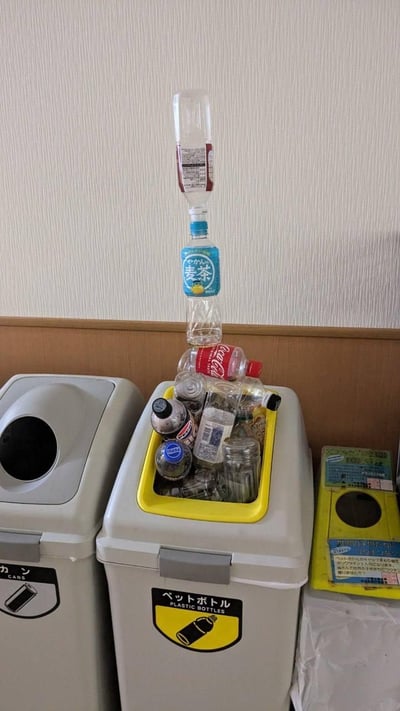 日本垃圾分类场景实拍：画面中央是一个黄色内圈的塑料瓶专用垃圾桶，桶内塞满了各种饮料瓶，包括透明瓶、棕色瓶和红色可口可乐瓶。最引人注目的是桶口处有一组塑料瓶被垂直堆叠起来，形成约30厘米高的“塔状结构”，其中一个蓝色标签的“麦茶”瓶清晰可见。垃圾桶左侧还有一个灰色的普通垃圾桶，背景是米白色墙面和棕色木质腰线，右侧贴有日文垃圾分类说明贴纸。整个场景展现了日常生活中有趣的垃圾分类瞬间，塑料瓶的堆叠方式显得既随意又富有喜感。