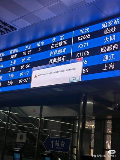 火车站电子显示屏上显示着多趟列车的始发站、终到站、开点和站台信息，如K2665次开往临汾、K371次开往大同、K1155次开往成都西等，状态均为“在此候车”。画面中央突兀地弹出一个错误提示窗口，内容为“NVIDIA GeForce GTX 750 Ti 设备是不可移动的，无法被虚拟或出租。”，形成了现实场景与电脑硬件错误提示的幽默混搭效果。