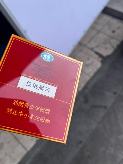 这是一张上海烟草集团上海卷烟厂出品的展示用香烟包装盒实拍图，包装盒采用红底金边的经典样式，盒面标注了该烤烟型香烟的焦油量11mg、烟气烟碱量1.1mg、烟气一氧化碳量11mg等参数，盒身贴有白色的“仅供展示”贴纸，底部印有“劝阻青少年吸烟 禁止中小学生吸烟”的控烟警示标语，拍摄背景为室外的砖石地面与建筑边缘。