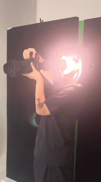 图片中显示一名穿着黑色上衣的人正在用相机拍照，脸部被强烈的逆光效果笼罩，形成明亮的光晕。人物手臂上有一个纹身图案，背景是黑色幕布，右侧有绿色元素。整体画面呈现出专业摄影的工作场景，逆光效果营造出戏剧性的光影对比。