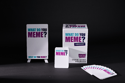 图片展示了WHAT DO YOU MEME?卡牌游戏的中国首发版本产品套装。画面中有一个白色展示牌、一个主包装盒、一张单独卡牌和一叠散开的游戏卡片，所有物品均以白色为底色，印有蓝紫色的"WHAT DO YOU MEME?"标题文字。主包装盒上还有"中国首发版本"的中文标识，以及"一款轻松鬼畜口超有梗的表情包聚会桌游"的产品描述。整体构图简洁，以黑色背景突出白色卡牌产品。