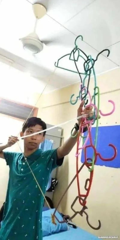一名身穿绿色带白点T恤的男生在宿舍内，用多个彩色塑料衣架捆绑组合成一把简易弓箭，左手持弓右手拉弦，神情专注地摆出射箭姿势。背景可见宿舍常见的天花板吊扇、窗户和蓝色墙面装饰，展现了大学生活中脑洞大开的DIY创意。
