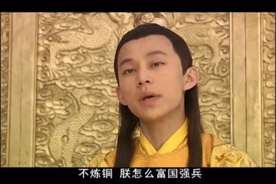 这是一张经过字幕修改的电视剧剧照截图，画面中何炅饰演的明朝正德皇帝身着明黄色龙袍、头戴皇冠，表情严肃，正开口说话，背景是雕刻着龙纹与云纹的金色墙面。图下方的字幕为“不炼铜 朕怎么富国强兵”，这里运用了谐音梗，“炼铜”谐音“恋童”，以皇帝的口吻说出这句荒诞的台词，制造出反差搞笑的效果。