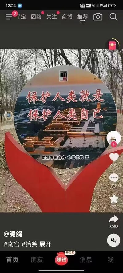 这是一张竖屏的短视频平台屏幕截图，画面核心是一个户外设置的宣传标语牌：圆形的标语牌被红色的V形支架托举，背景是光秃的树木和泥土地面，标语牌上印有南宫当地建筑的夜景图，上方有“南宫”字样，中间的宣传语为“保护人类就是保护人类自己”，这句标语存在明显的逻辑重复，属于文案乌龙，标语牌下方标注了发布单位“南宫市创森办 市城管局 宣”。截图的界面显示这是在一款带有“赚钱”导航栏的短视频APP中，发布者为@鸽鸽，内容带有#南宫 #搞笑 标签，界面右侧显示该视频的互动数据：点赞583、评论212、收藏27、转发3088，底部导航栏有“首页”“朋友”“赚钱”“消息”“我”五个选项，还有“开宝箱”的按钮。