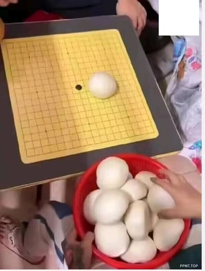 这张图片呈现了一个充满趣味的恶搞场景：传统的围棋棋盘上，没有摆放常规的黑白棋子，而是用一个白面馒头替代棋子放置在棋盘上，棋盘下方的红色塑料盆里装满了同款白面馒头，显然是要将这些馒头当作下棋的“棋子”来使用。画面中可以看到多只手，其中一只手正放在装馒头的盆上，似乎准备取用“棋子”，整体场景将日常食物与传统围棋玩法结合，通过强烈的反差感制造出搞笑的效果，充满生活趣味。