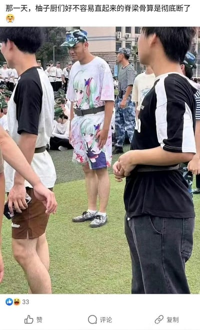 图片展示了军训场地的场景，一群身着军训服装或便装的年轻人聚集在草坪上。其中一名男子格外显眼，他穿着印有绿色长发动漫女性角色的白色T恤和同款图案的短裤，脚踩运动鞋，戴着迷彩帽，双手自然下垂站立。图片上方配有文字“那一天，柚子厨们好不容易直起来的脊梁骨算是彻底断了😂”，通过“柚子厨”（对特定动漫角色“柚子”的粉丝昵称）的自嘲，幽默地表现出该男子穿着夸张动漫“痛衣”出现在严肃军训场合，让其他同好感到尴尬又好笑的场景。