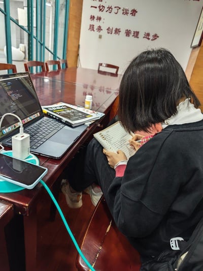 这是一张在室内自习空间拍摄的照片，一位短发女生坐在长桌旁，正低头专注地在笔记本上书写记录。桌上摆放着打开的苹果笔记本电脑（屏幕显示代码界面）、正在充电的手机、努比亚充电器、书本和小药瓶，背景墙上写着"一切为了读者"以及"服务 创新 管理 进步"的标语，整体场景传递出认真学习的专注氛围，没有双关、谐音梗或暗示内容。