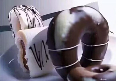 这是一张利用视觉错位效果拍摄的创意美食照片，画面中的巧克力淋面蛋糕卷与巧克力甜甜圈，通过巧妙的摆放位置，让观者第一眼产生误判，仿佛看到了一个带有白色绑带的深色U形物体，仔细观察才能发现是两种甜点的错位组合，营造出充满趣味的视觉反差效果。