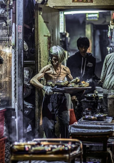 这张纪实摄影作品展现了香港街头一家狭小拥挤的熟食店后厨场景：一位身形瘦削、肌肉线条清晰的赤裸上身的师傅，戴着隔热手套，端着一盘冒着热气的食物，神情专注又带着疲惫地在充满油烟与蒸汽的环境中穿梭。他的皮肤和衣物上都沾染着油烟痕迹，背景里还有其他工作人员在灶台前忙碌，整个空间被厨具、蒸汽填满，充满了浓厚的市井烟火气，也直观体现出街头餐饮从业者的辛劳日常。