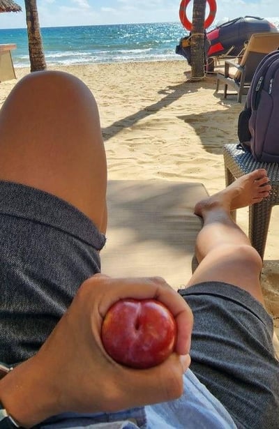 这是一张在海滩度假场景下拍摄的创意视觉错位照片。画面中，拍摄者躺在沙滩躺椅上，一只手握着一颗色泽红润的李子，巧妙利用拍摄视角，将李子放置在膝盖前方的位置，让李子看起来仿佛是从膝盖上“生长”出来的凸起部分，制造出荒诞又有趣的视觉错觉。背景是阳光照耀下的金色沙滩与波光粼粼的蓝色大海，一旁还有沙滩椅、救生圈等度假设施，整体场景充满了轻松惬意的度假氛围，这个无心或有意的视角巧合让普通的日常瞬间变得趣味十足。