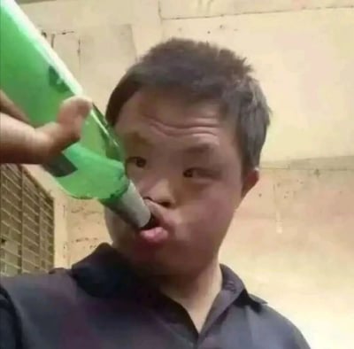 这是一张利用视觉错位效果制作的搞怪图片，画面中的真人做出嘟嘴的动作，从特定拍摄角度看，绿色玻璃酒瓶的瓶口恰好与人物的嘴部位置重合，营造出仿佛酒瓶瓶口被塞入口中的怪异视觉效果。人物眉头微皱，面部因嘟嘴动作显得滑稽，背景是砖墙结构的半室内环境，整体画面充满趣味，常被用作搞笑表情包在网络传播。