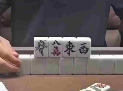 这是一张实拍的麻将对局场景图，画面靠前位置特意摆放了四张麻将牌，从左到右依次为幺鸡、八万、东风、西风，利用四张牌名称的谐音组合成脏话“鸡巴东西”，属于趣味谐音玩梗内容。画面左侧可见玩家摆弄麻将的手部，下方是码放整齐的其他麻将牌，右下角还露出了另外两张麻将的局部，背景模糊，整体是生活化的玩麻将场景。