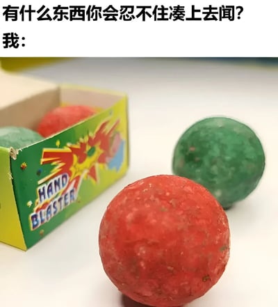 图片上方文字提问：'有什么东西你会忍不住凑上去闻？'，下方紧接着'我：'并配上一张童年玩具的照片。照片中展示了一个黄绿相间的玩具盒，盒身上印有'HAND BLASTER'字样和爆炸图案，盒内及盒外分别放着红色和绿色的颗粒状球体。这张图通过幽默的方式，用童年常见的HAND BLASTER玩具球作为答案，勾起人们对这类玩具的怀旧记忆，尤其是可能曾出于好奇凑上去闻过的经历。