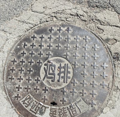 这是一张实拍的路面井盖静态图，圆形铸铁井盖安装在开裂的水泥路面上，井盖表面布满用于防滑的十字形凸起，中间圆形区域的文字因磨损等原因，视觉上看起来和食品名称“鸡排”完全一致，井盖下方边缘印有铸造厂相关的标识文字，已有部分磨损，该巧合形成了趣味视觉梗。