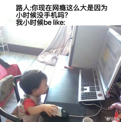 这是一张顶部配文的搞笑网络梗图，顶部文字内容为“路人:你现在网瘾这么大是因为小时候没手机吗？我小时候be like:”，下方是实拍的生活场景：一名年幼的小女孩坐在椅子上，一只手握着台式电脑的鼠标，脸凑得非常靠近桌面，正十分专注地盯着面前的老式台式电脑屏幕，画面生动回应了配文的问题，表明自己从小就已经接触电脑、沉迷上网，网瘾是从小养成的，以此调侃反驳“现在网瘾大是小时候没有手机导致”的说法，充满趣味。
