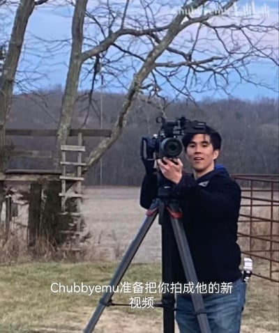 图片中显示一位亚裔男性正专注地操作着三脚架上的专业摄像机，背景是户外的自然场景，有光秃秃的树木、梯子和围栏，看起来像是在进行外景拍摄工作。图片底部有文字'Chubbyemu准备把你拍成他的新视频'，表明这可能是视频创作者Chubbyemu在为新作品进行拍摄准备。