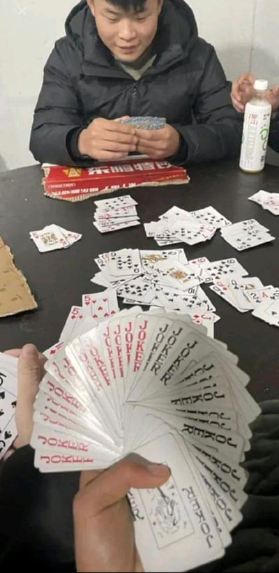 图片展示了一个令人惊讶的扑克牌游戏场景。一名穿着黑色外套的男子坐在桌子旁，面前散落着许多扑克牌。最引人注目的是前景中有人手中拿着一大把Joker牌，数量之多令人难以置信，形成了强烈的视觉冲击和幽默感。桌子上还可以看到东鹏特饮的红色包装盒和一瓶饮料，暗示这可能是一场长时间的牌局。整个场景营造出一种荒诞而搞笑的氛围，因为在正常的扑克牌游戏中不可能出现如此多的Joker牌。