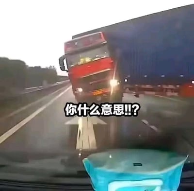 这是一张从车辆前挡风玻璃（行车记录仪视角）拍摄的雨天高速公路场景图，画面里一辆红色车头蓝色货箱的重型大货车位置倾斜，车头正对着拍摄车辆方向，视觉上给人一种大货车即将迎面撞过来的强烈冲击力，画面中部叠加了黑色白边的文字“你什么意思!!?”，结合发布背景，这张图用来传递愚人节被整蛊后，面对离谱状况的错愕、质问的情绪。