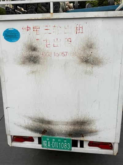 这是一张快递行业电动三轮车的后部实拍照片，白色的车厢面板上有多处污渍和磨损痕迹，面板上喷涂有红色的“纳星三轮出租”“电出租”字样，还有部分被污渍遮挡的联系电话数字；左下角贴有一个蓝色圆形标识。车厢底部是绿色的快递行业专用车牌，牌号为皖A·DN1083，车牌上方标注“快递行业”，车牌两侧装有红色的尾灯。整体车辆外观显示出长期使用的陈旧感，是城市配送场景中常见的运输工具。