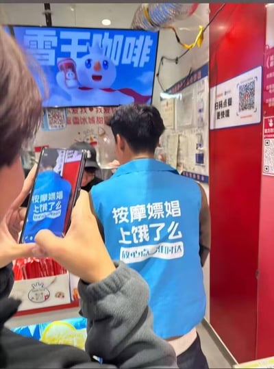 在蜜雪冰城门店内，一名穿着蓝色饿了么马甲的男子背对镜头站在柜台前，旁边有人正用手机拍摄他的后背。马甲上的官方标语被恶意修改，上方替换为低俗违规的“按摩嫖娼 上饿了么”，下方保留了饿了么原本的“放心点 准时达”，制造出荒诞又低俗的恶搞反差效果。
该内容通过篡改外卖平台官方马甲的合规标语，替换成违规低俗内容，形成违背品牌形象的整活梗。