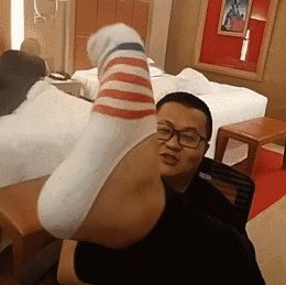 这是一张动态GIF搞怪图，场景位于酒店房间内，一名戴眼镜的男子坐在椅子上，将自己的脚伸到镜头前方，借助视觉错位的拍摄手法，让脚看起来像是直接长在自己头部的位置，制造出荒诞滑稽的视觉效果，男子面带笑意，整体氛围搞笑逗趣。