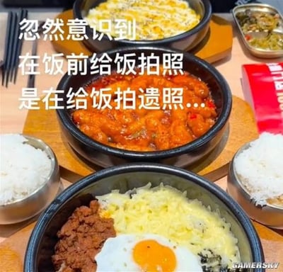 这是一张结合美食实拍的搞笑梗图，画面主体是摆放在餐桌上的多份韩式料理，包括盛放在石锅内的辣炒年糕、搭配了芝士、肉末与溏心蛋的拌饭、芝士类餐食，两侧摆放着金属碗装的白米饭，右上角还有小份配菜和红色包装物品。画面上叠加了三行白色醒目的文字，用夸张的类比调侃了当下很多人用餐前先给食物拍照分享的常见行为，将食物被食用前的留影类比为遗照，形成反差感的笑点。