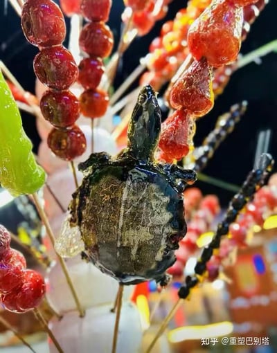 图片展示了一堆色彩鲜艳的糖葫芦串，主体是传统的红色山楂糖葫芦，表面裹着晶莹剔透的糖衣，在灯光下泛着光泽。在这些常规山楂串中间，穿插着一个造型奇特的黑色乌龟形状糖葫芦，乌龟的头部和龟壳轮廓清晰，同样覆盖着糖衣，显得格外突兀和有趣。背景为深色，突出糖葫芦的鲜亮色彩，整体给人一种传统小吃与创意造型结合的新奇感。