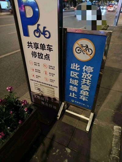 夜间街道旁并排竖立着两块内容矛盾的共享单车指示牌。左侧为白色底的“共享单车停放点”指示牌，顶部有蓝色P字及自行车图标，下方列有多项使用规定和管理公示栏信息；右侧为蓝色底的禁止停放指示牌，顶部有红色圆圈加斜杠的禁止自行车图案，下方文字明确写着“此区域禁止停放共享单车”。两块相邻的指示牌形成鲜明对比，引发对停放规则的困惑。