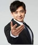 图片中是一位穿着黑色西装、白色衬衫和深色领带的男性，他面带微笑，右手自然伸出，掌心朝向镜头，背景为简洁的浅灰色纯色背景，整体给人正式且友好的感觉。
