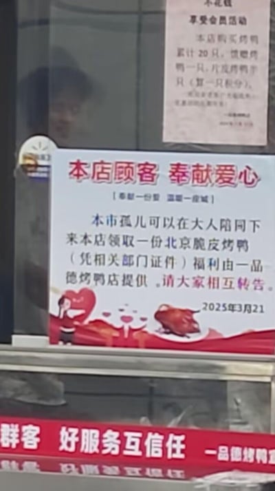 这是一张线下一品德烤鸭店玻璃的实拍照片，核心是蓝底的爱心公益告示，标题为「本店顾客 奉献爱心」，副标题为「奉献一份爱 温暖一座城」，内容显示本市孤儿可在大人陪同下，凭相关部门证件到店领取一份由该店提供的北京脆皮烤鸭，落款2025年3月21日，并呼吁大家相互转告。告示下方有红色店铺标语「群客 好服务互信任 一品德烤鸭」，玻璃上方还贴有会员活动告示，告示搭配可爱的卡通爱心形象，整体传递出商家暖心的公益氛围。