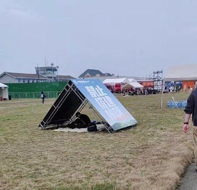 一片开阔的草地上，一个写着“禁止搭建帐篷”的大型三角指示牌自己倒塌了，形成了一个类似帐篷的三角形结构，下面还压着一些物品和可能的人。背景中可以看到多个帐篷和建筑物，远处还有人在活动，形成了一种讽刺的场景。
