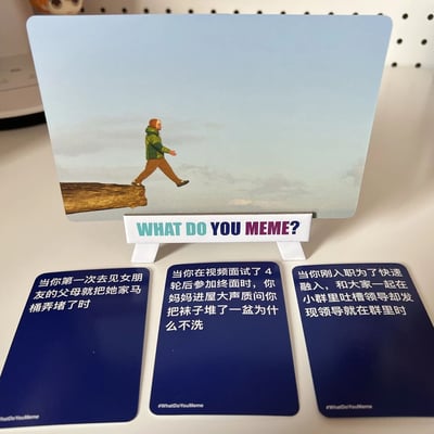 图片展示了热门 meme 卡牌游戏《What Do You Meme?》的实体卡牌实拍场景。中央立着一张主图卡，画面中一名穿着绿色外套、棕色裤子的人正迈步走在悬空的悬崖边缘，脚下是云海缭绕的高空，营造出惊险又荒诞的氛围。主图卡下方由白色支架支撑，支架正面印有游戏标题“WHAT DO YOU MEME?”（彩色字体）。主图卡前方平摆着三张蓝色 caption 卡牌，每张卡牌上用白色文字描述了不同的尴尬生活场景：
1. “当你第一次去见女朋友的父母就把她家马桶弄堵了时”
2. “当你在视频面试时，4轮后参加终面时，你妈妈进屋大声质问你把袜子堆了一盆为什么不洗”
3. “当你刚入职为了快速融入，和大家一起在小群里吐槽领导却发现领导就在群里时”
卡牌右下角均带有“#WhatDoYouMeme”的标签。整体构图清晰，通过主图的惊险画面与下方尴尬文字的反差制造幽默效果，展现了该游戏“用 meme 卡片搭配搞笑文字”的核心玩法。