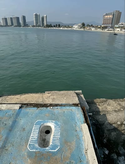 一张海边风景照，前景是一个蓝色的水泥结构，上面有一个类似蹲式厕所的洞口，后方是宽阔的海面和远处的城市天际线，包括多栋高楼大厦和一个正在建设中的建筑。海滩上有一些遮阳伞和休闲设施，整体呈现出一种城市海滨的景象。照片的视角从厕所洞口位置望向海面，形成了一种奇特的视觉对比。