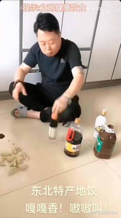 这是一段搞笑短视频的截图，画面中一名男子盘腿坐在地上，手持毛笔，旁边摆放着类似酱油、料酒的瓶装液体，地上的泥状物体被调侃为“东北特产地饺”，配文“嘎嘎香！嗷嗷叫！”，顶部还有“提示:文明健康饮食”的文字。内容玩了“地饺”的谐音梗，模仿东北特产宣传语的语气，用荒诞的整活方式恶搞美食视频，制造出滑稽搞笑的反差效果，同时顶部的提示语和整活内容形成反向调侃。