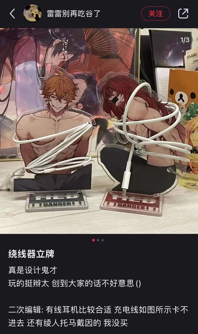 社交媒体用户“雷雷别再吃谷了”发布的帖子截图，展示了两个动漫男性角色的亚克力立牌被创意性地用作绕线器，白色数据线缠绕在角色身体上。立牌底座印有“I DANGER!!”字样，背景可见其他动漫海报和周边。帖子配文称“绕线器立牌 真是设计鬼才”，并补充说明有线耳机更适合使用，充电线卡不进去，还提到绫人、托马、戴因等角色的立牌未购买。整体内容幽默有趣，展现了二次元爱好者的创意收纳方式。
