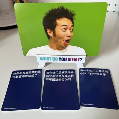 图片展示了一款名为"What Do You Meme?"的中文卡牌游戏实物。画面中央是一张立起来的主卡牌，上面是一位表情极度夸张的惊讶男子特写，背景为纯绿色。卡牌下方有三个蓝色的 caption 卡牌，分别写着："当你朋友圈里的炮王突然宣布要结婚了"、"当你进门后发现你的两个基佬室友在进行不可描述的行为时"和"当一个四川人骂他的小孩'你个龟儿子'"。所有卡牌上都有"#WhatDoYouMeme"的水印。