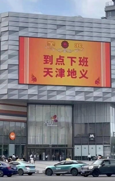 这是一张拍摄于天津街头的实景照片，画面主体是某商业综合体的户外电子大屏，大屏以橙红色为背景，显示着醒目的标语“到点下班 天津地义”，巧妙运用谐音梗，将成语“天经地义”中的“经”替换为天津的简称“津”，调侃天津人到点下班是理所当然的事，贴合天津悠闲的生活氛围，充满趣味。大屏顶部还配有“津”“沽”字样，呼应天津“津沽”的别称。画面下方是城市街道，停着几辆汽车（包括天津特色的青绿色出租车），还有不少行人在商场入口附近活动，呈现出日常的城市街景。