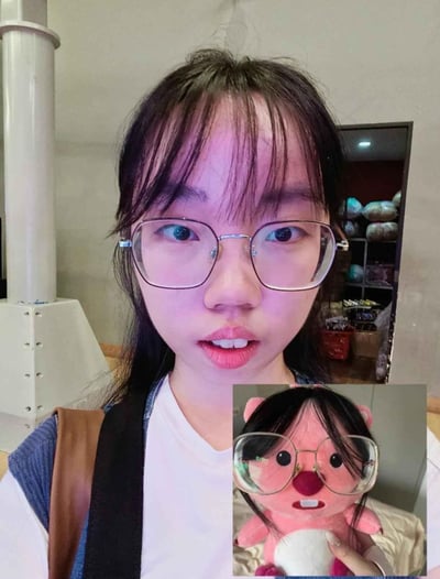 图片主体是一位戴眼镜的女性直视镜头，表情略显惊讶。右下角有一个小图，显示一个粉色毛绒玩具戴着相似款式的眼镜，表情呆萌。两者在眼镜款式和面部表情上有明显相似之处，形成有趣的对比效果。背景中可以看到室内环境，包括白色柱子和放置物品的架子。