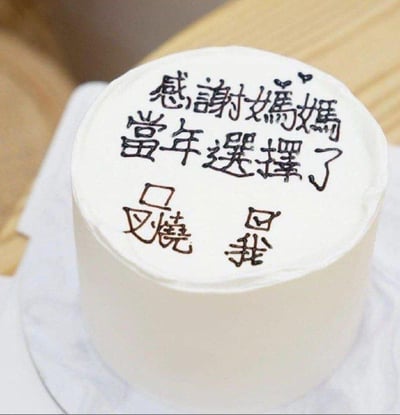 这是一款白色奶油裱面的圆形创意蛋糕，蛋糕表面用黑色可食用色素手写了经过繁体转简体的文字：“感谢妈妈当年选择了我”，其中“我”字被设计成模仿“叉烧”的字形结构，玩了粤语地区的经典谐音梗——在粤语俗语里有“生叉烧都好过生你”的抱怨式说法，意思是觉得孩子还不如叉烧，这款蛋糕反用这个梗，以自嘲又温情的幽默方式，表达对妈妈选择养育自己的感谢，既有趣又充满亲情温度。