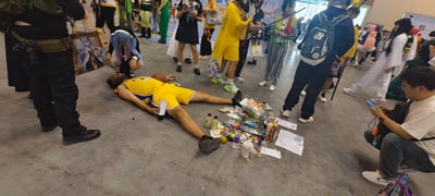 这是在国内漫展现场的趣味整活场景：一名身着湖人24号黄色球衣的男子平躺在地面上，身边散落着篮球、饮料瓶、纸张、零食袋等物品，模仿“倒地昏迷”的状态。周围聚集了多名不同风格的coser，包括修女装扮、古风装扮、JK装扮的参与者，有的蹲在旁边围观，有的站着拍照，现场氛围轻松欢乐，是漫展参与者自发的趣味互动，没有双关、谐音梗或隐含暗示，纯粹是现场搞活制造笑点。
