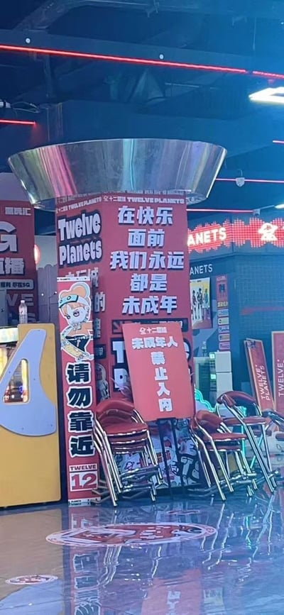 图片展示了名为'Twelve Planets'的电玩城内景。中央矗立着红色立柱，上面用白色大字写着'在快乐面前我们永远都是未成年人'，立柱旁另有一块红色指示牌写着'未成年人禁止入内'，形成有趣的反差萌。立柱左侧有卡通女孩形象，手持物品，旁边标注'请勿靠近'。周围可见堆叠的红色座椅、黄色电玩设备及天花板上的霓虹灯条，整体氛围充满年轻活力。标语巧妙地将成年人追求快乐的心态比作'未成年'，与'未成年人禁止入内'的规定形成幽默对比，引发观众共鸣。